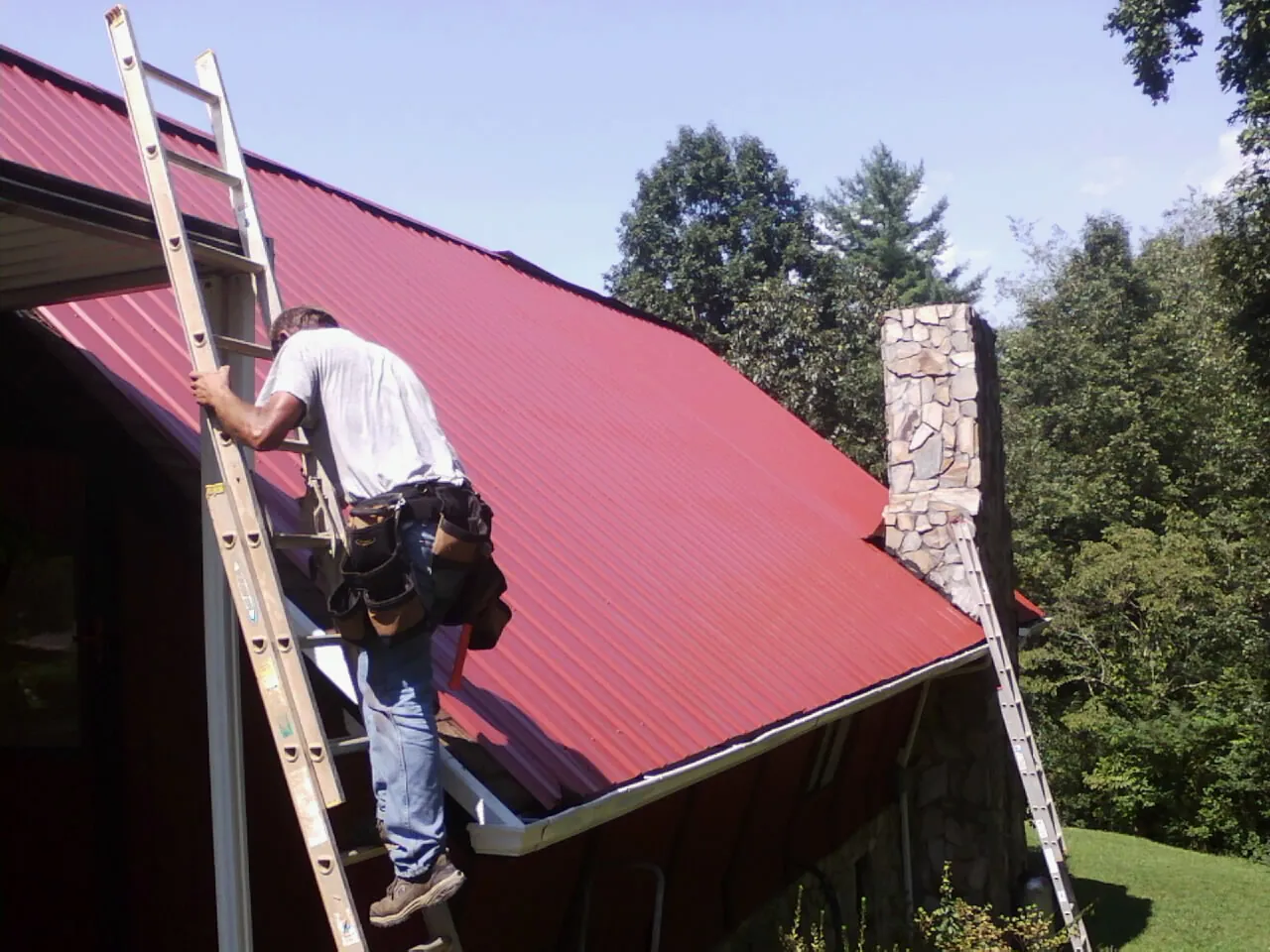 Roofer on a ladder beside a red metal roof and stone chimney — Soffit & Fascia Repair by Roofing Brothers in North Plains